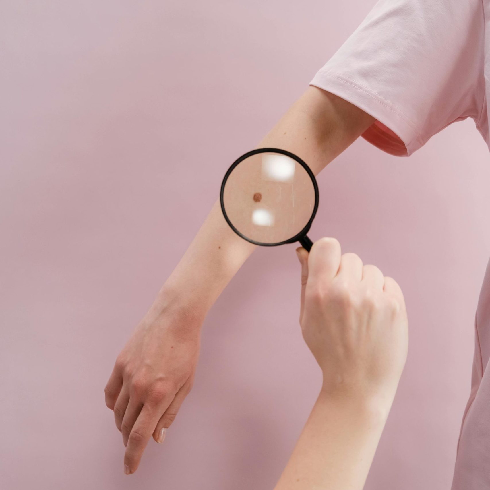 Close-up of a dermatologist examining skin with a magnifier on a pink background.